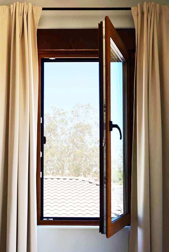 European-style tilt-and-turn window partially open, framed by light curtains, showcasing a view of a tiled roof and trees outside.