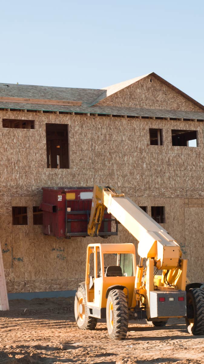 Construction site featuring a telehandler lifting a red material container towards a new building framed with plywood, highlighting modern construction processes.