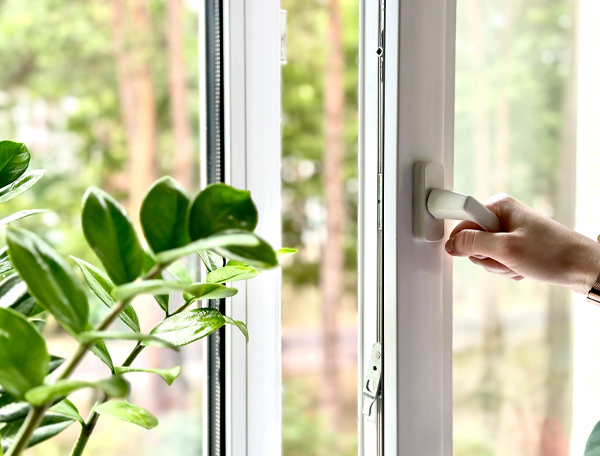 Hand opening a modern window with a sleek handle, surrounded by green indoor plants, emphasizing European design and functionality in window solutions.