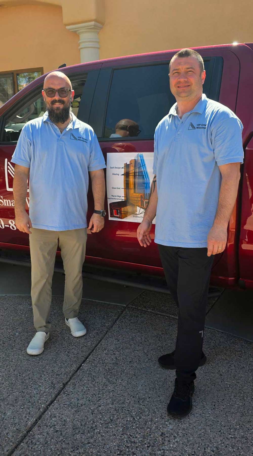 Two men wearing light blue polo shirts with the VIP Smart Windows logo, standing beside a red vehicle displaying a poster with window design details, in an outdoor setting.